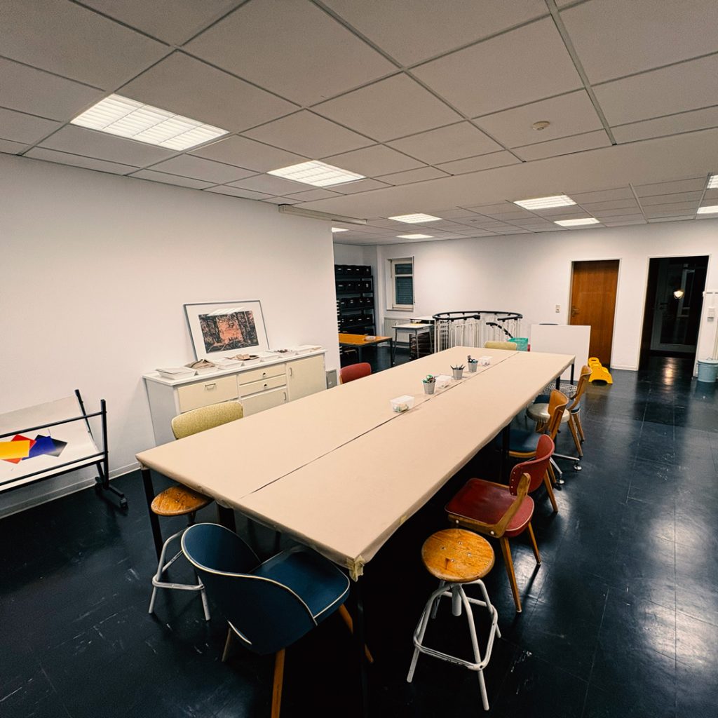 Einblick in die Malschule: Großer Tisch mit bunten Stühlen zum Arbeiten.