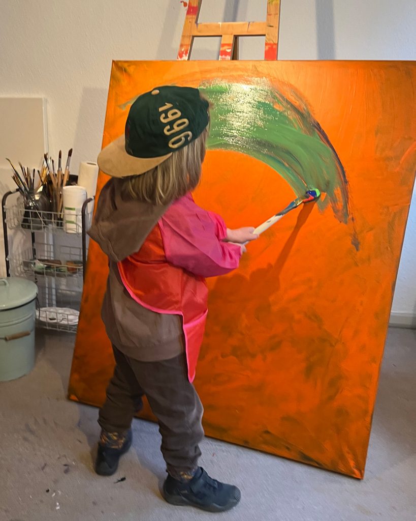 Ein Kind vor einer großen bunten Leinwand mit einem Pinsel in der Hand.